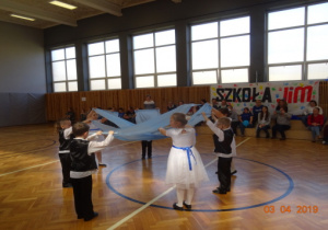 Chłopcy ubrani na galowo i dziewczęta w białych sukienkach tańczą ze skrzyżowanymi niebieskimi szarfami.